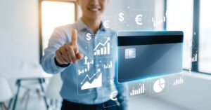Pessoa sorridente em escritório moderno aponta para cartão de crédito digital azul e branco em destaque, cercado por ícones de dinheiro e gráficos financeiros.